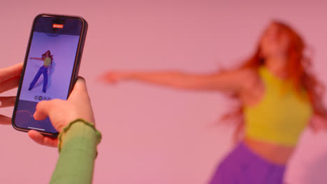 Studio-Shot-Of-Woman-Taking-Photo-Of-Friend-Dancing-On-Mobile-Phone-Against-Pink-Background-6